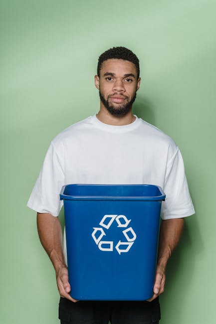 A person wearing a white long-sleeve shirt is placing a cardboard sheet into a blue recycling bin that features a white recycling symbol on the front. Next to the blue bin, there is a smaller white wa