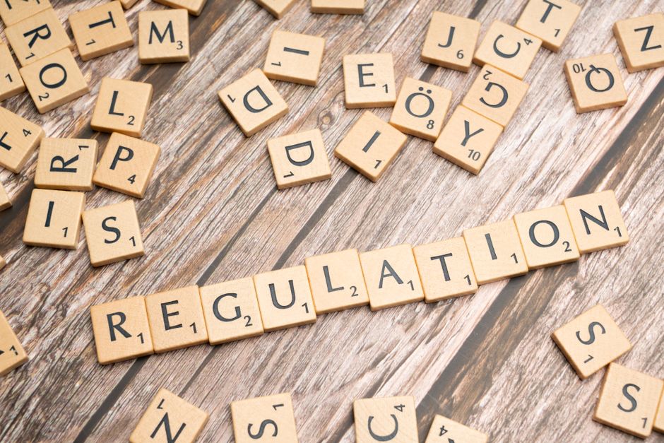 A collection of light wood Scrabble tiles with black letters and point values scattered across a wooden surface, some tiles aligned to spell the word 'REGULATION' in the center. The scattered tiles in